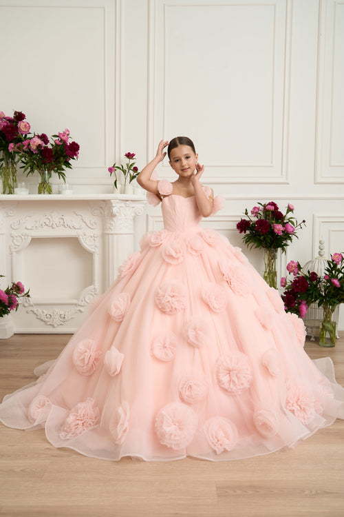Young girl in a pink floral dress standing in a room with decorative flowers and a fireplace.